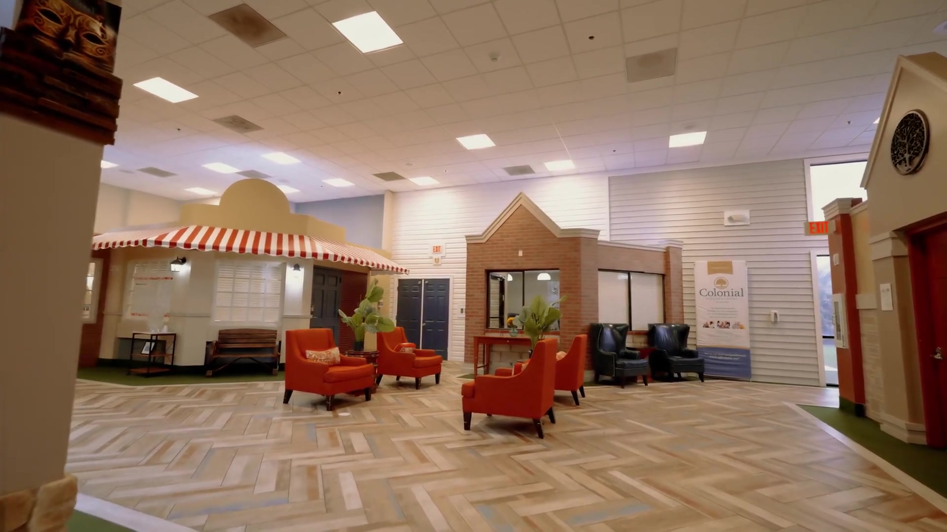 Welcoming lobby seating area with orange armchairs, herringbone wood floors, and striped awning at Colonial Gardens Residences assisted living community in Lauderhill, Florida